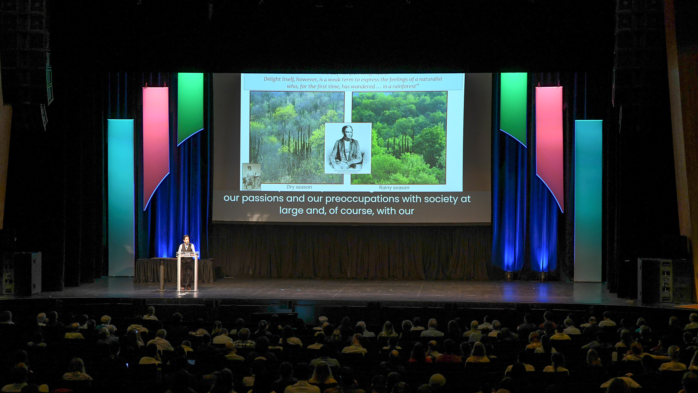 A plenary speaker presents before an audience of hundreds in an auditorium with a power point slideshow aid.