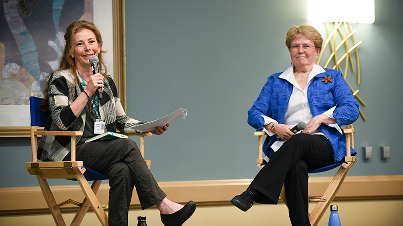 A session with two speakers seated in director's chairs. The background features a painting and a wall mounted sconce. 