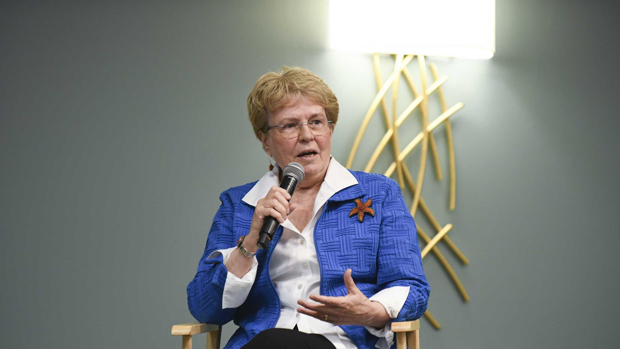 A presenter speaks to the audience from a director's chair.