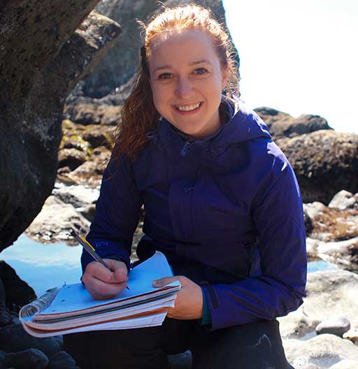 A younger ecologist takes notes by the rocky sea shore.