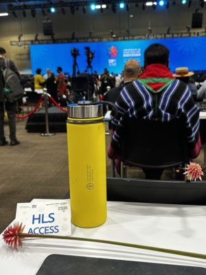 Close-up of a yellow water bottle, an HLS access badge, and a small flower on a table, with attendees and cameras in the background at COP16 in Cali, Colombia.