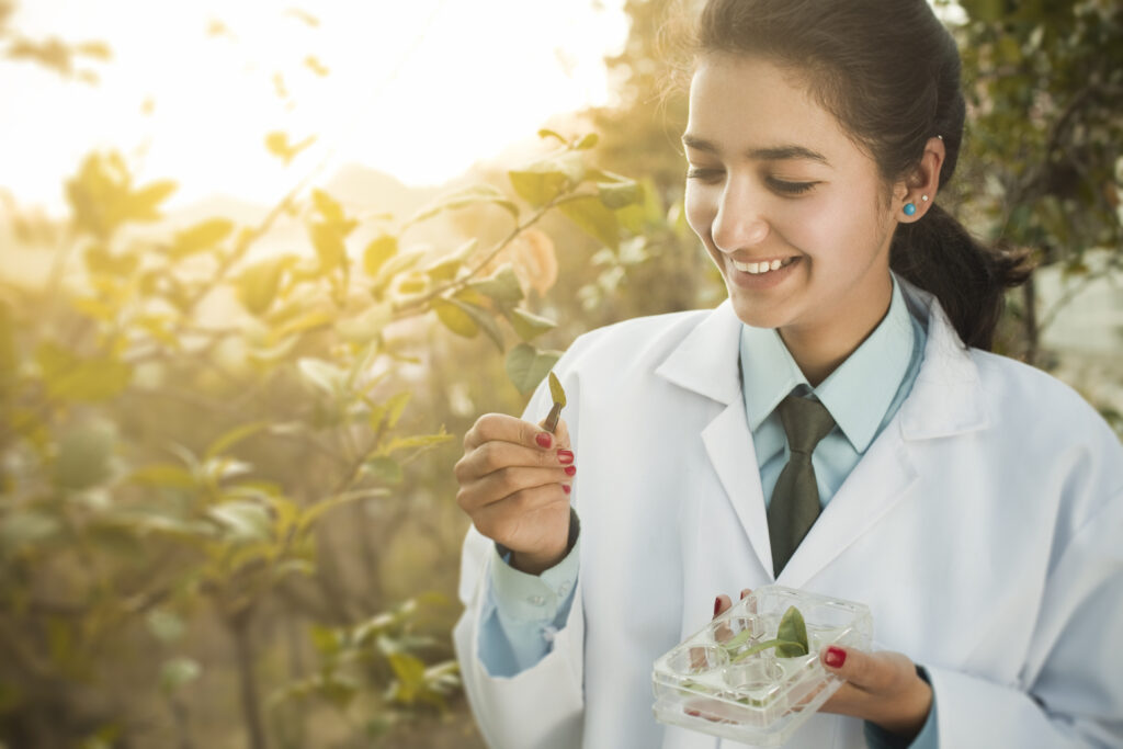 a young woman working as a researcher smiles as she examines a small plant sample in what appears to be a greenhouse