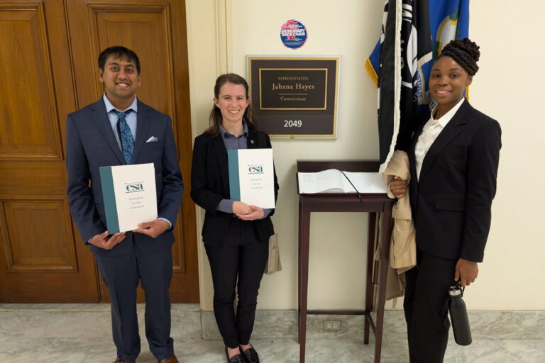 Three people in suits stand in front of an office door with flags and a plaque