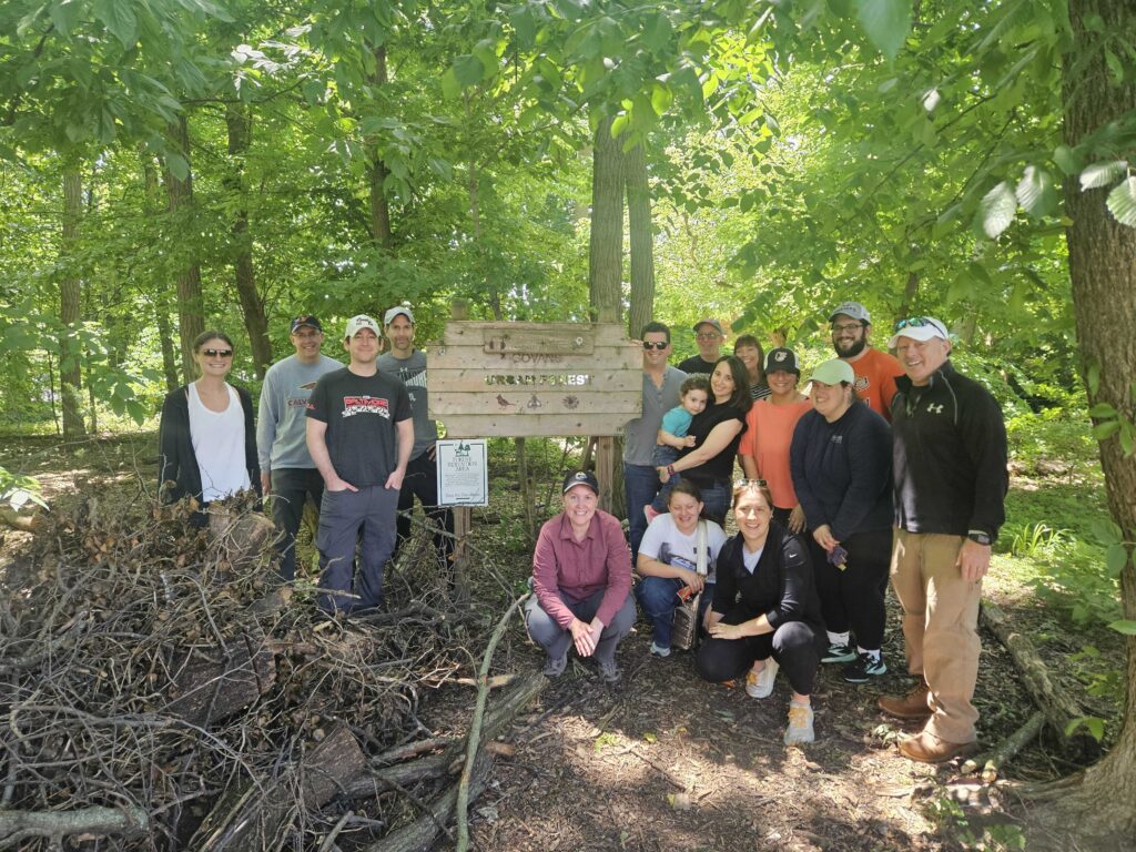 A group of people stands around a sign in a wooded setting.