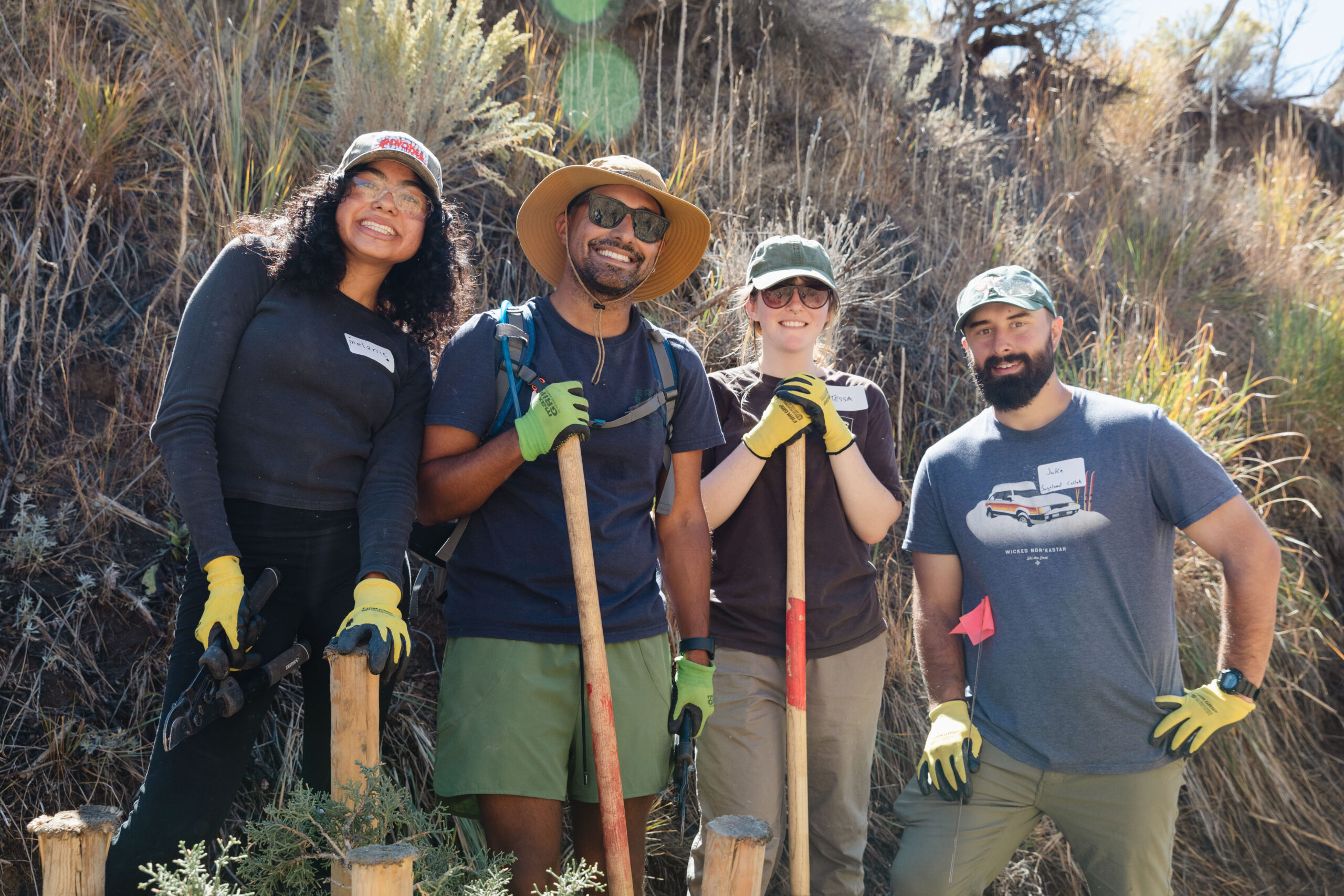Four people wearing tshirts and work gloves stand with hand tools in front of a sagebrush-covered slope.