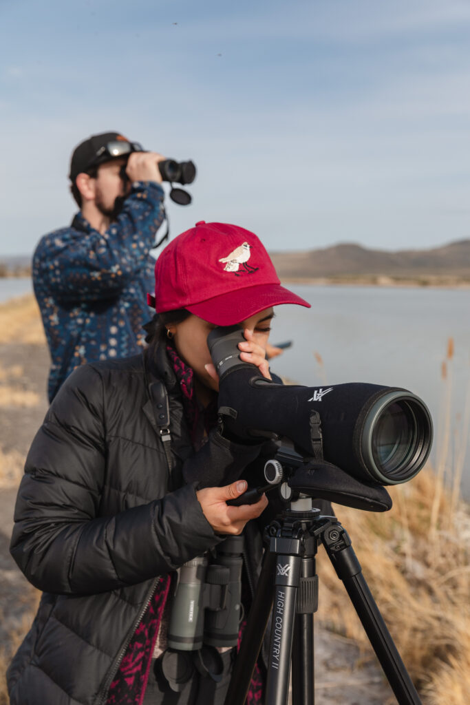Two people look through binoculars and a spotting scope on the edge of a lake.