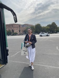 A smiling woman wearing a lanyard and holding a tote bag stands next to a bus.