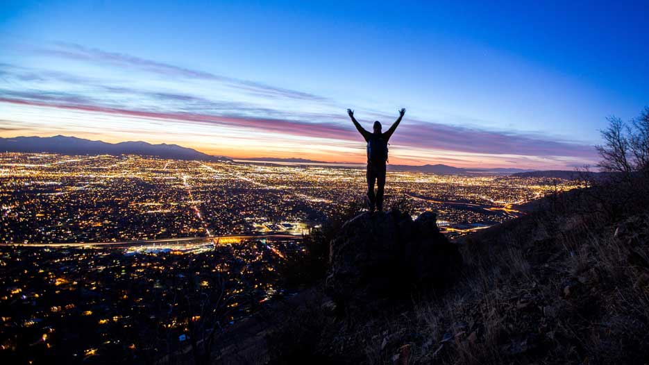 A person stands atop a cliff overlooking salt lake city at night with hands raised to convey enormity or conquering of personal struggles.
