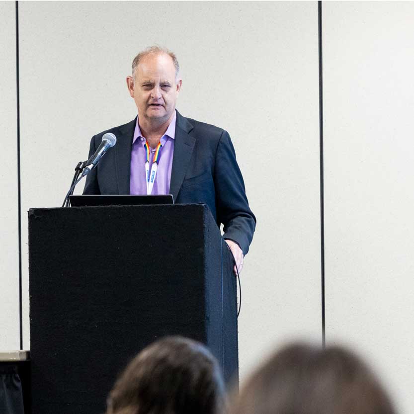 A presenter speaks into a microphone at a podium before an audience. 