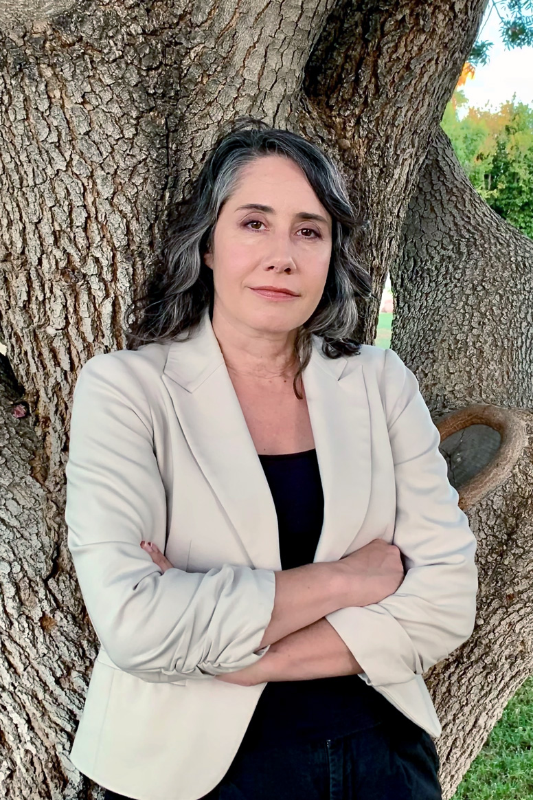 Diane Pataki stands before a thick tree, arms folded with a gentle smile. 