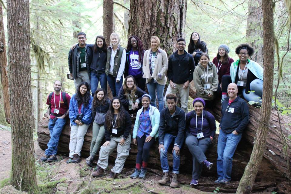 A group of SEEDS participants stand before and lean against tall trees in a forest. 