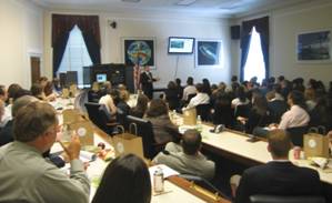A presenter speaks before a table of people.