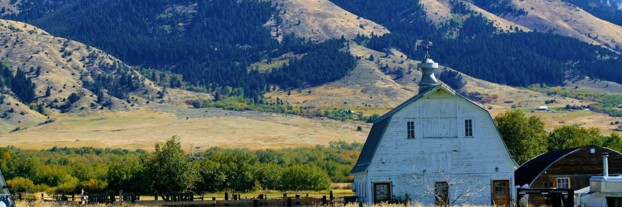 A barn next to a farmhouse with a mountainous background with fields in the foreground.