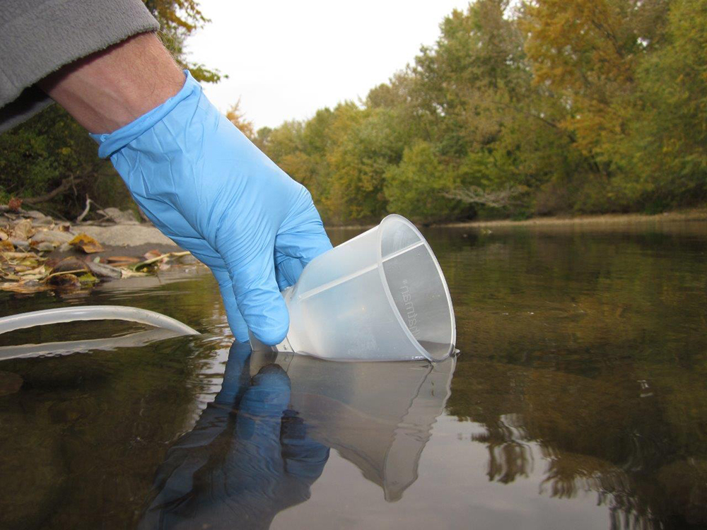 A gloved hand scoops river water with a sampling cup